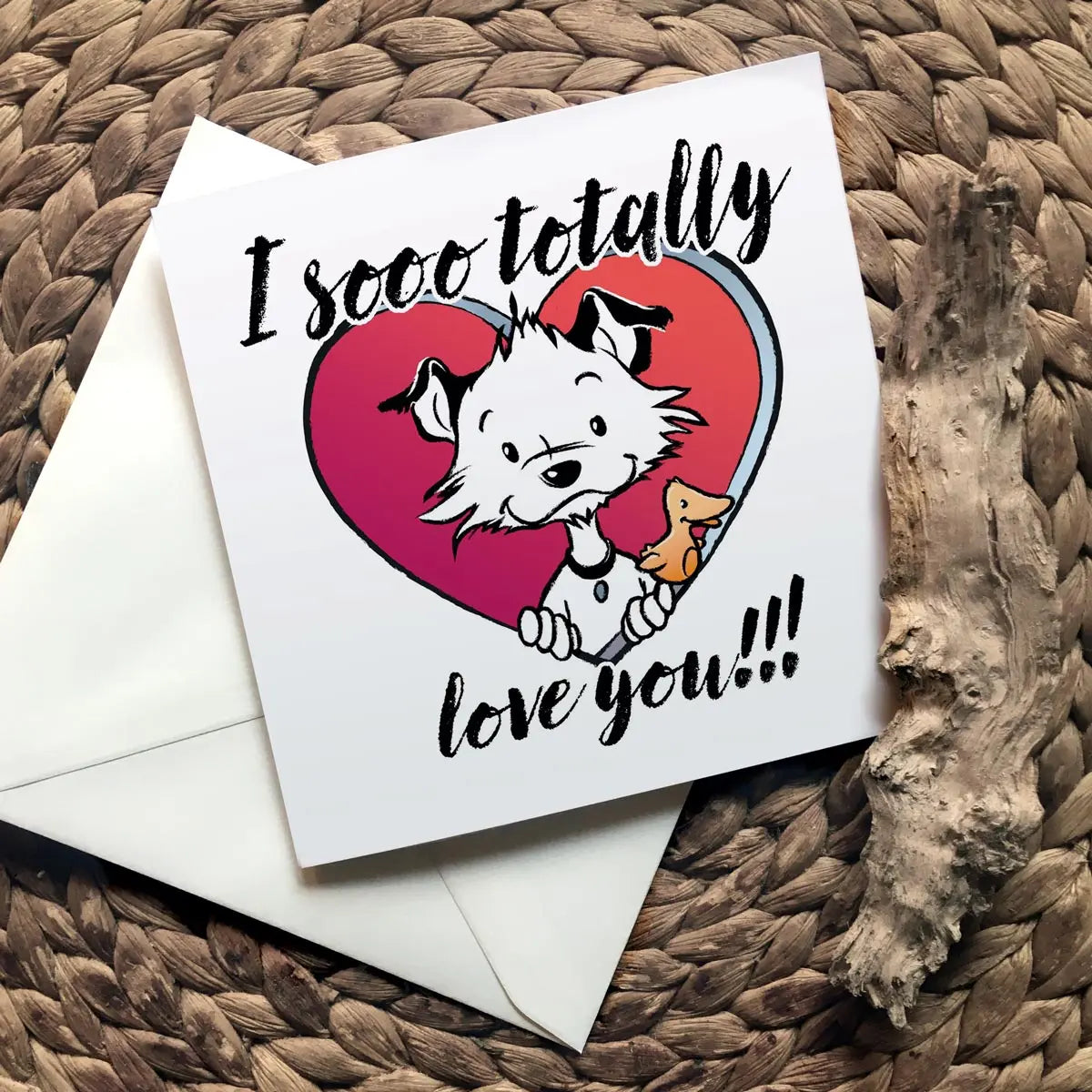 Image shows a square Little Hunterman card showing him and Flynn looking out of a big red heart with large text saying: I sooo totally love you!!! With closed card and envelope on a natural surface with one of Hunter's wooden sticks.