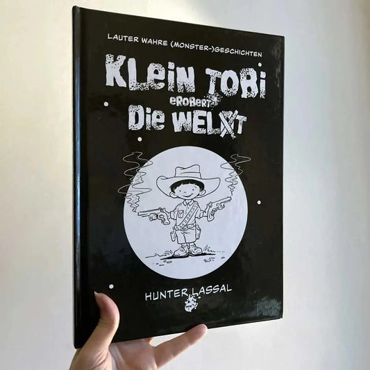 This image shows a hand holding the hardcover version of "Klein Tobi erobert die Welt: lauter wahre (Monster-)Geschichten" by Hunter Lassal against a light background. The front of the cover is facing the camera.