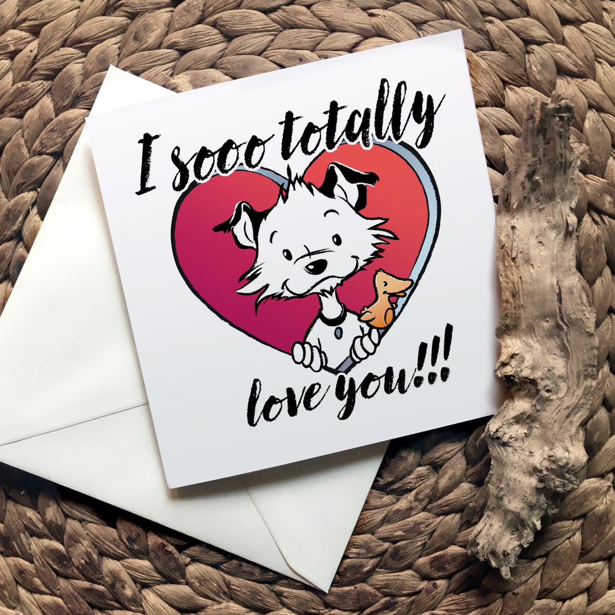 Image shows a square Little Hunterman card showing him and Flynn looking out of a big red heart with large text saying: I sooo totally love you!!! With closed card and envelope on a natural surface with one of Hunter's wooden sticks.