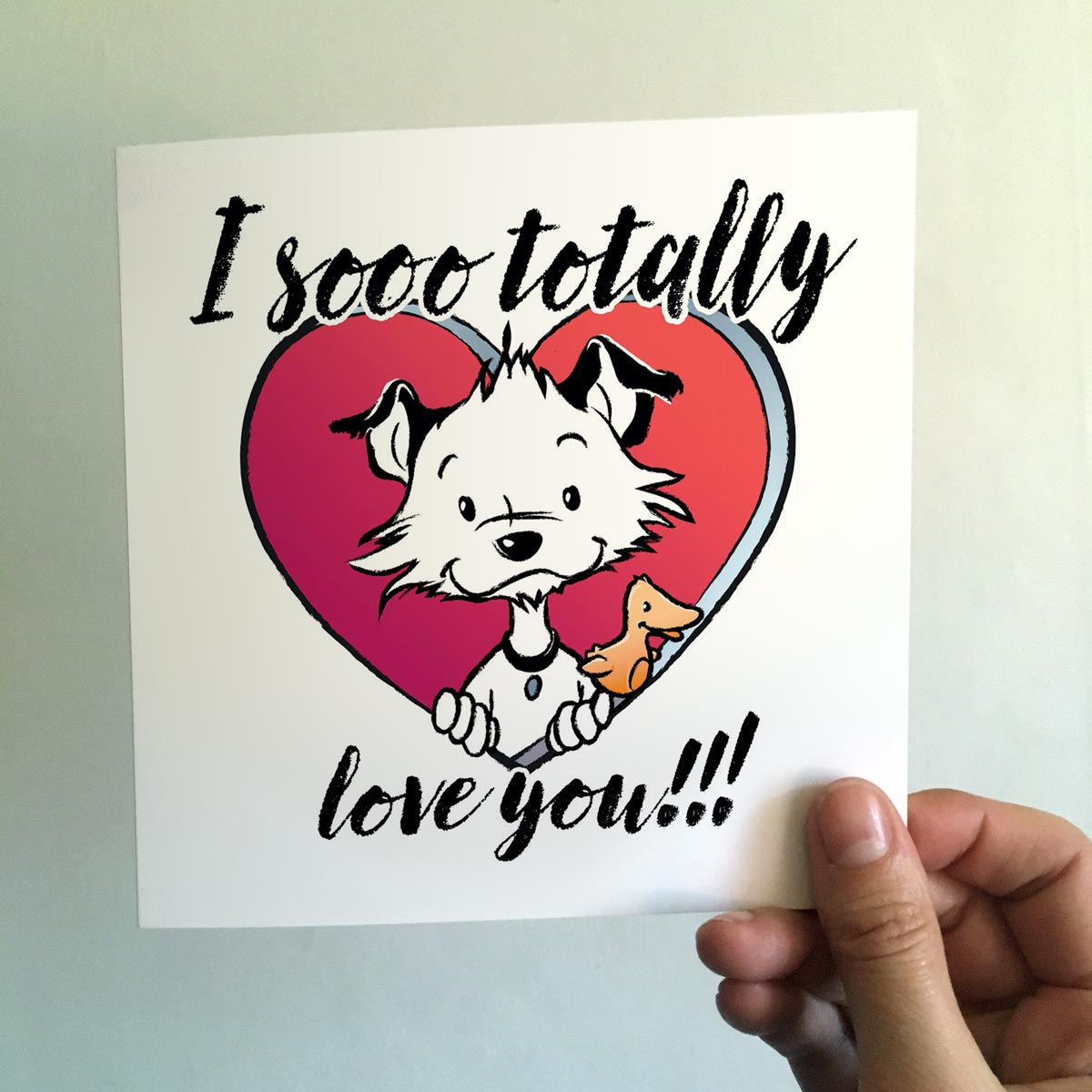 Image shows a square Little Hunterman card with him and Flynn looking out of a big red heart with large text saying: I sooo totally love you!!! With a hand holding the card agains a light background