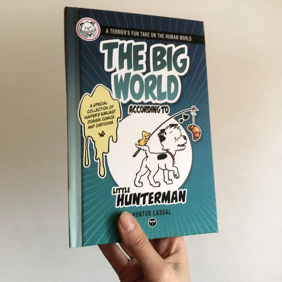 The Big World According to Little Hunterman, retailer version hardcover with black and white illustrations and creme colored paper. The image shows a hand holding the hardcover against a light wall – with the front cover towards the camera.