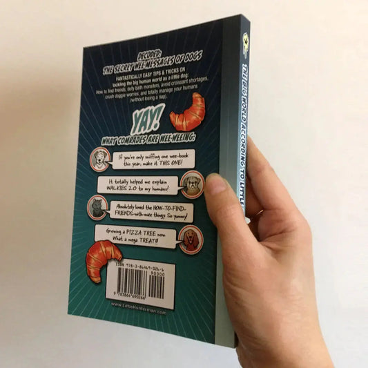 The Big World According to Little Hunterman by Hunter Lassal, retailer version paperback with black and white illustrations and creme colored paper. This image shows a hand holding the book against a light background with the back and spine facing forward.