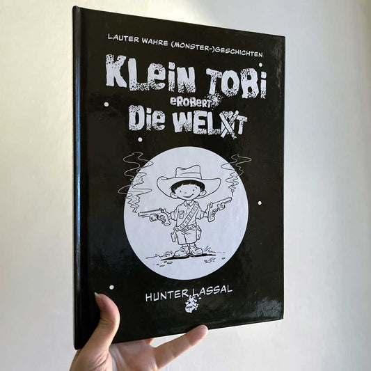This image shows a hand holding the hardcover version of "Klein Tobi erobert die Welt: lauter wahre (Monster-)Geschichten" by Hunter Lassal against a light background. The front of the cover is facing the camera. 