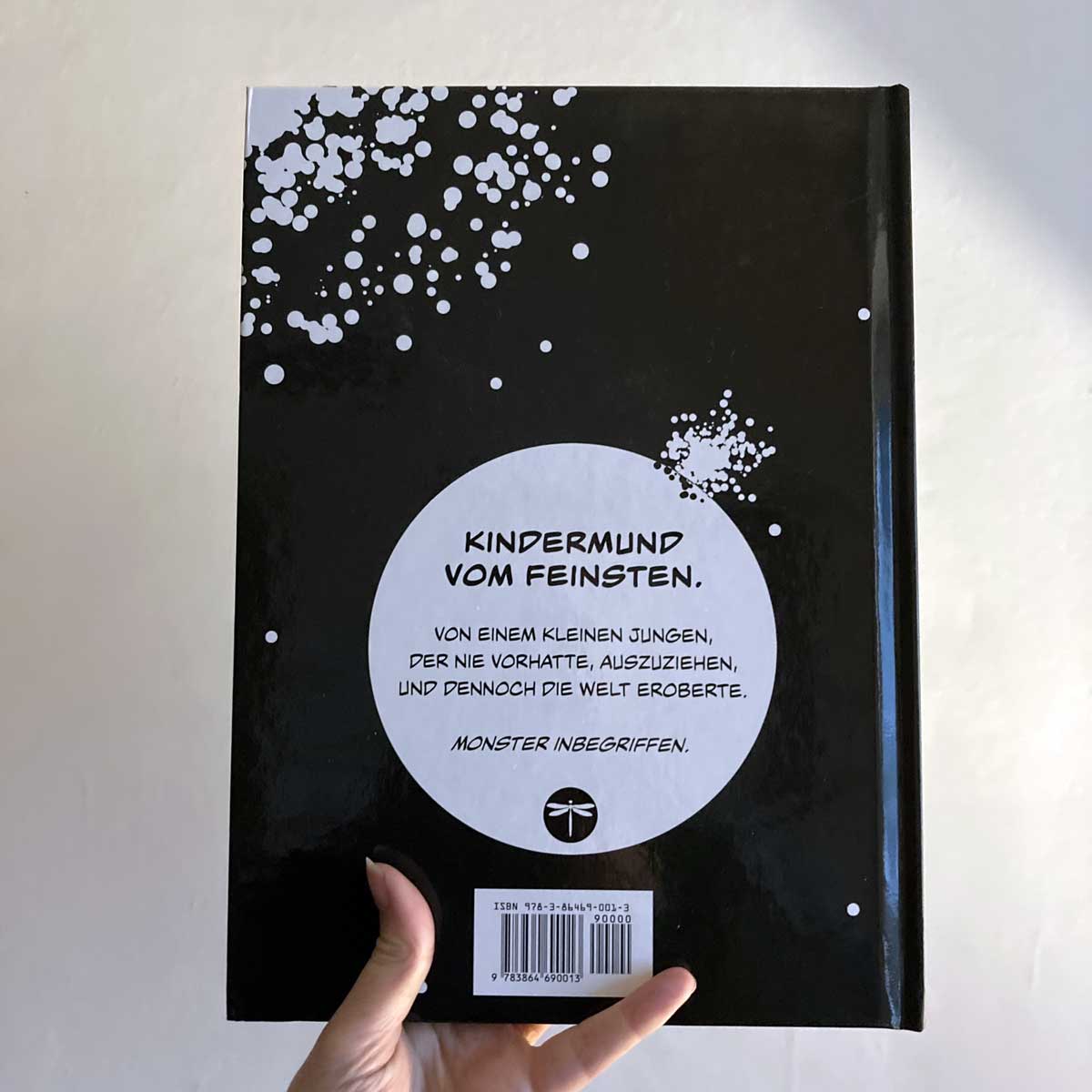 This image shows a hand holding the hardcover version of "Klein Tobi erobert die Welt: lauter wahre (Monster-)Geschichten" by Hunter Lassal against a light background. The back of the cover is facing the camera. 