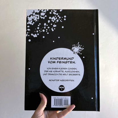 This image shows a hand holding the hardcover version of "Klein Tobi erobert die Welt: lauter wahre (Monster-)Geschichten" by Hunter Lassal against a light background. The back of the cover is facing the camera. 