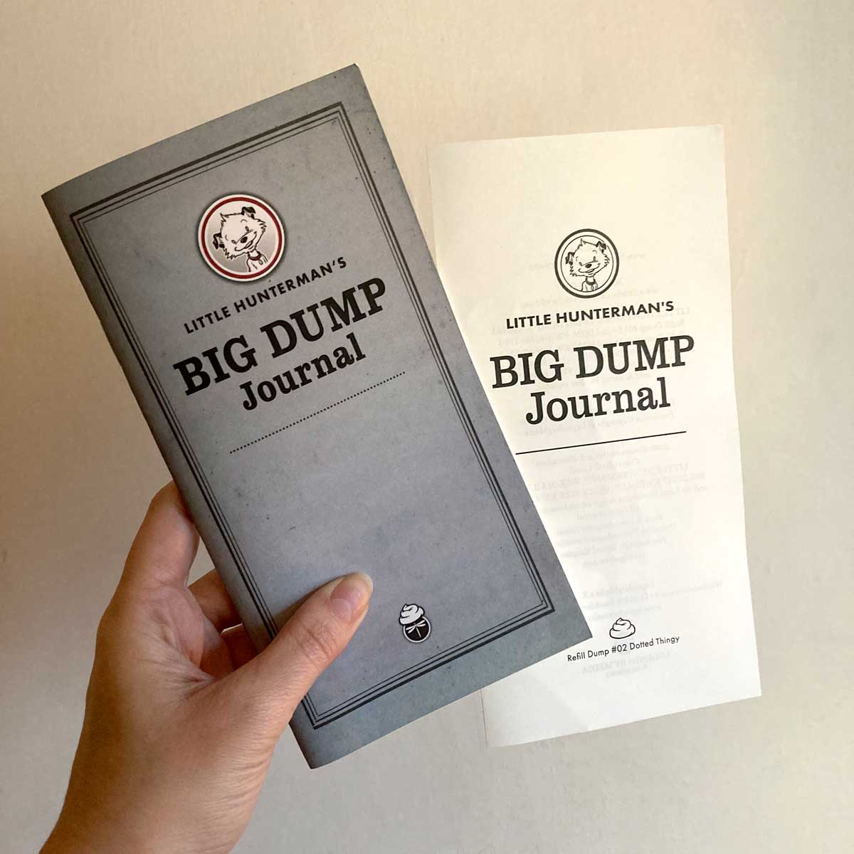This image shows a hand holding a journal against a light background. The journal, one of Little Hunterman's BIG DUMP Journals, is seen with a separate spread that contains the title page (and the imprint on the back). These pages come included with the journal despite not being stapled to it. 