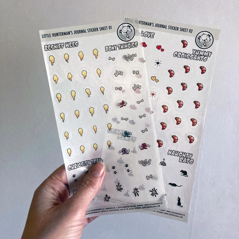 A hand holding one of Little Hunterman's Journal Sticker Sheets 01 and 02 up against a light background.