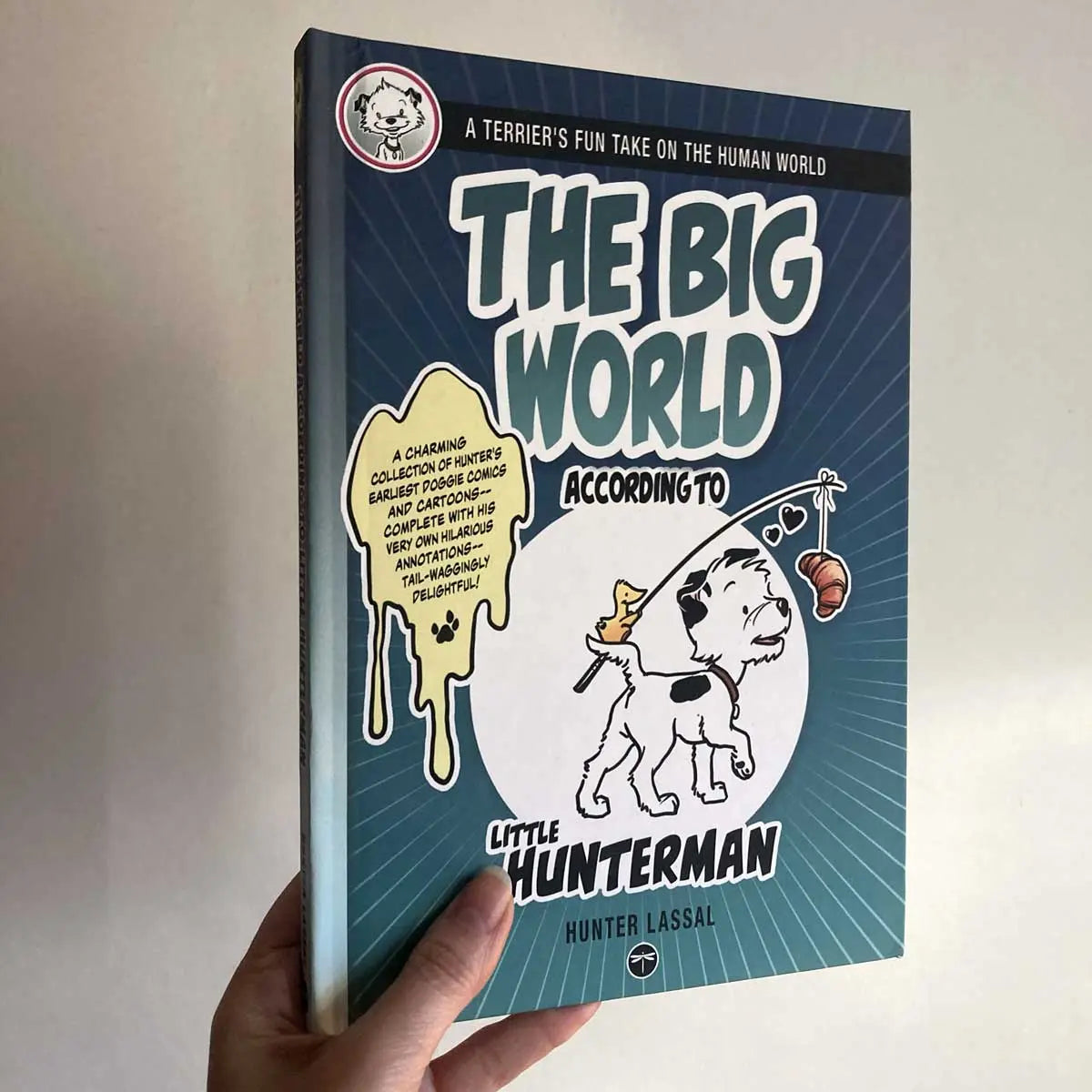 The Big World According to Little Hunterman by Hunter Lassal, premium hardcover with black and white illustrations on white, coated paper. The image shows a hand holding the hardcover against a light wall, with the front cover towards the camera. 