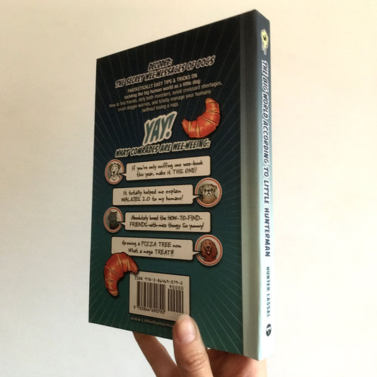 A hand holding The Big World According to Little Hunterman standard hardcover version against a light wall – with the back and spine towards the camera. 