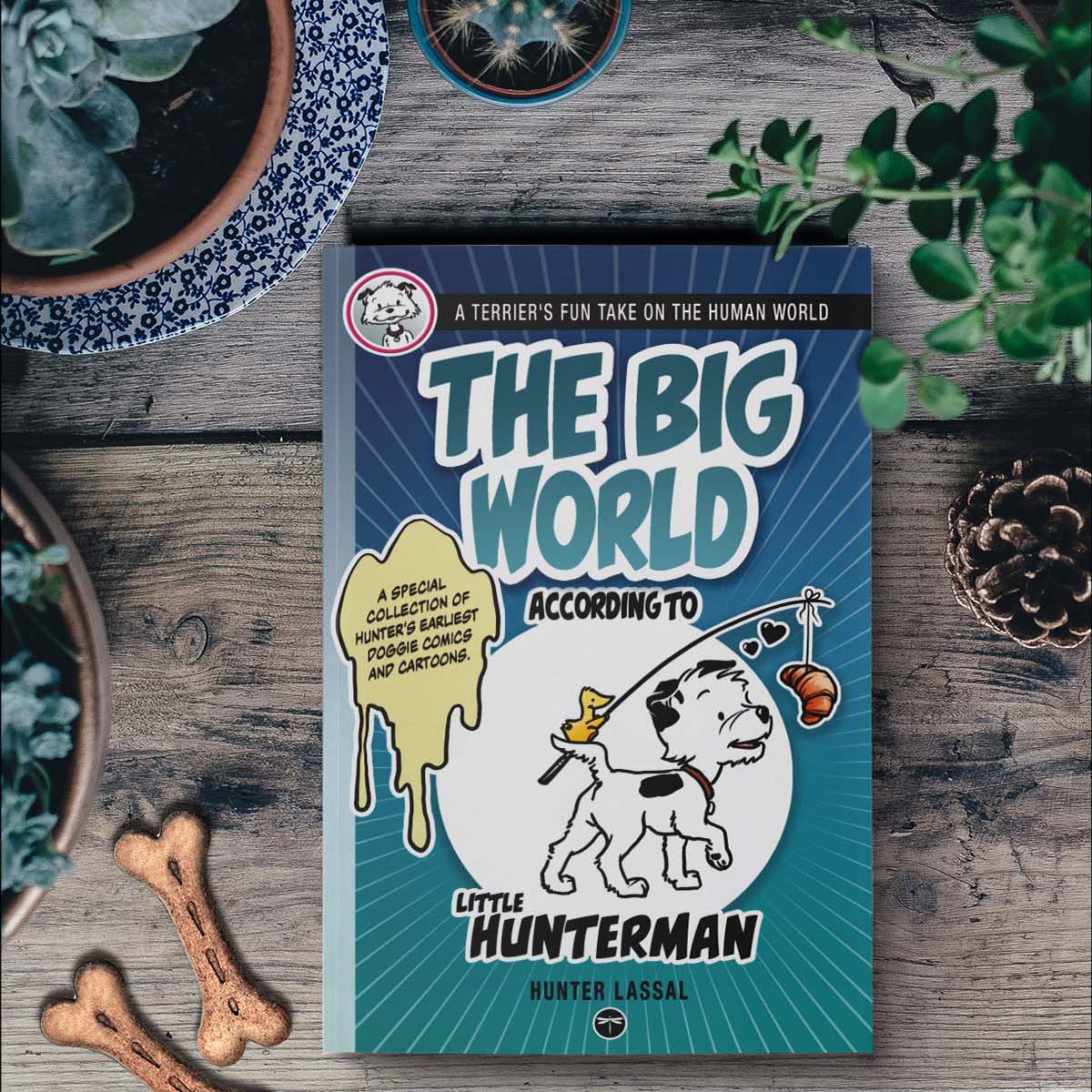 The Big World According to Little Hunterman, retailer version paperback with black and white illustrations and creme colored paper. This image shows the book on a table among plants and doggie treats.